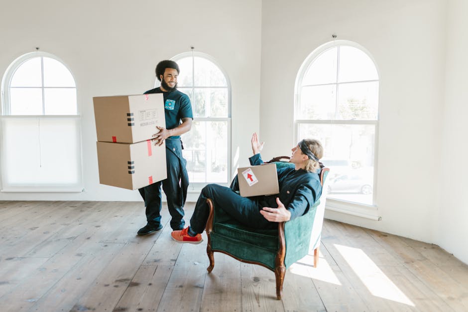 A professional removal service scene showing a man with dark curly hair and a beard, dressed in dark clothing, carrying two large cardboard boxes with red and black tape inside a bright, spacious room