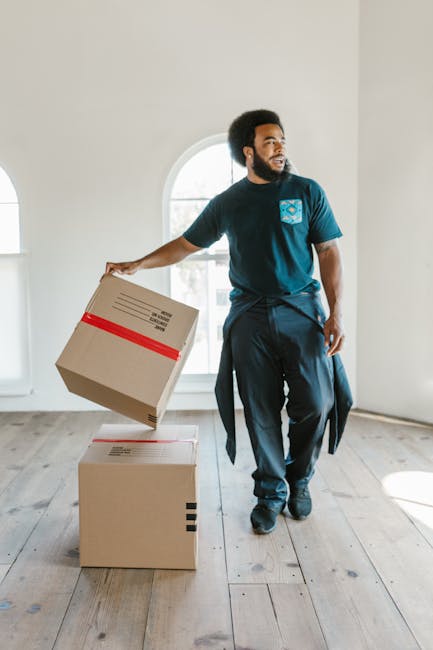 A professional removal service scene showing a man with dark curly hair and a beard, dressed in dark clothing, carrying two large cardboard boxes with red and black tape inside a bright, spacious room