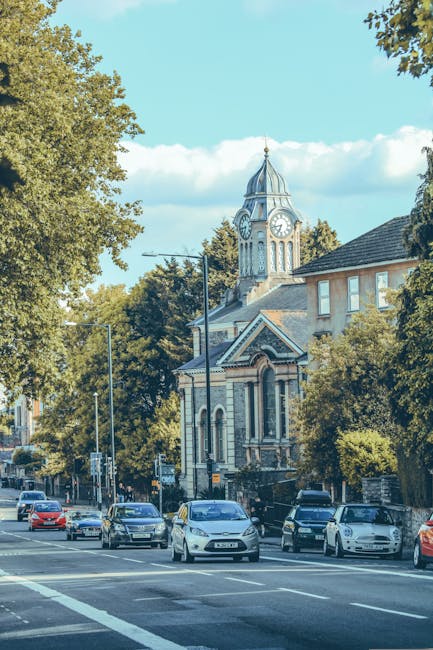 A street scene showing a row of parked cars along the pavement in front of a historic building with a clock tower and a domed roof, surrounded by lush green trees under a partly cloudy sky. The building appears to be a church or similar structure, with brick and stone façades. On the left side of the image, there are large leafy trees providing partial shade, with some traffic lights and street signs visible near the vehicles. The scene is well-lit, suggesting daytime. In the context of house removals, the image depicts an urban environment where furniture and boxes could be loaded onto moving vans, such as those operated by Man and Van Balham, to facilitate home relocation or professional furniture transport services. The arrangement indicates a typical setting for logistical planning during packing and loading processes for moving day.